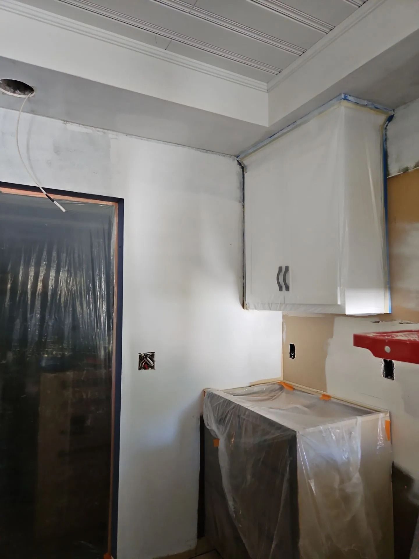 Kitchen renovation: white cabinets, covered appliances, unfinished walls, and door opening.