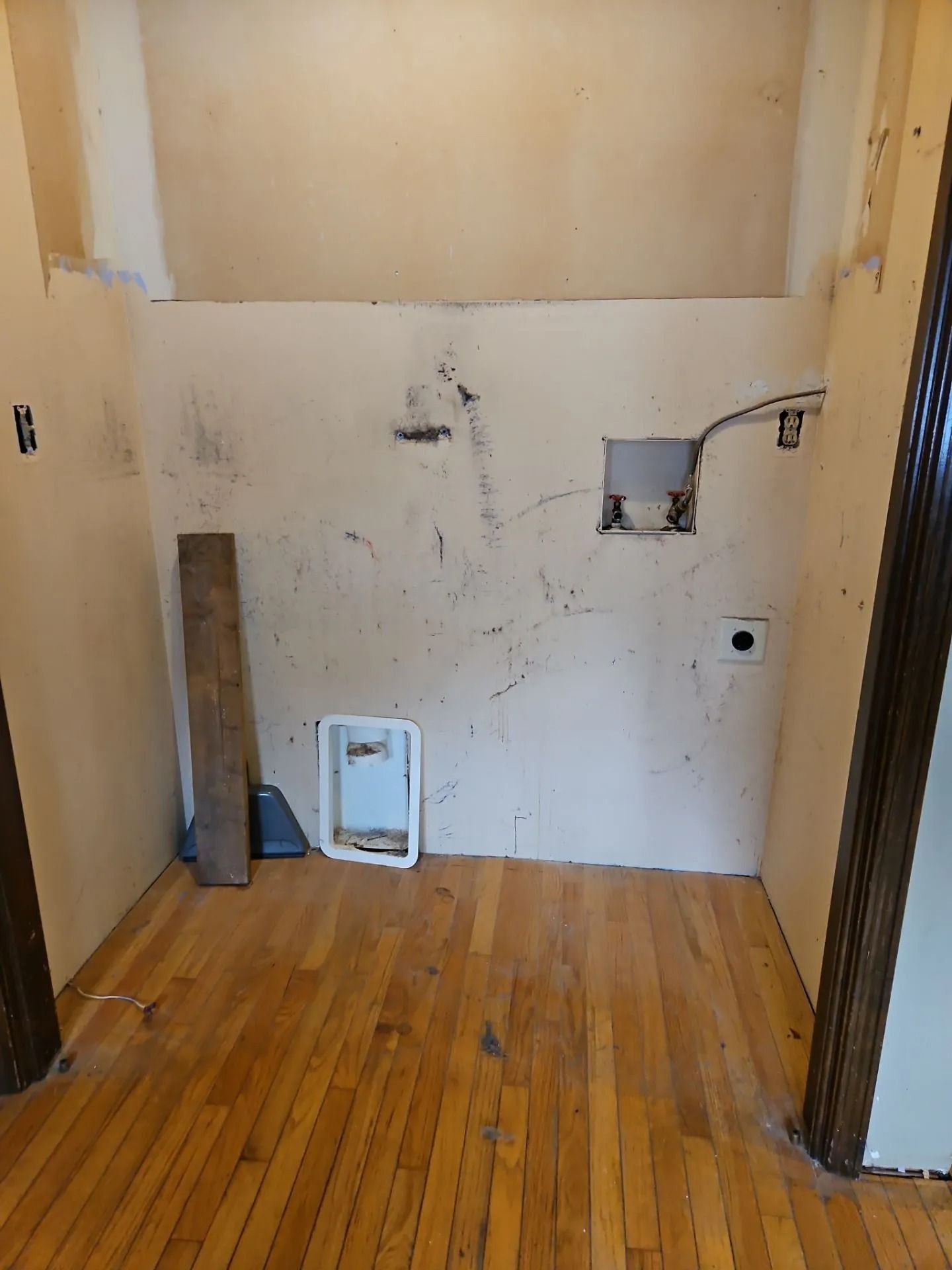 Empty laundry room with wood floor and stained walls. Electrical and plumbing are visible.