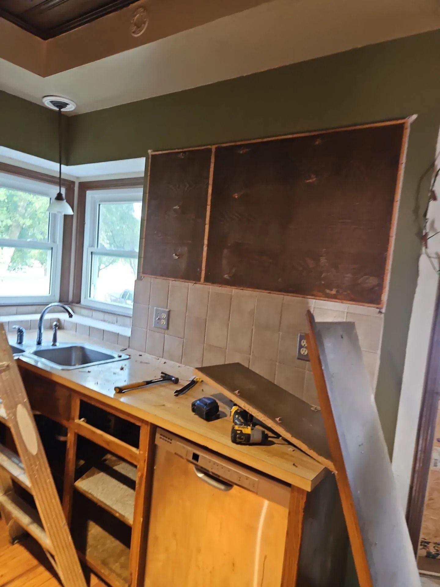 Kitchen renovation in progress, cabinet partially disassembled. Tile backsplash, olive green wall, bay window.
