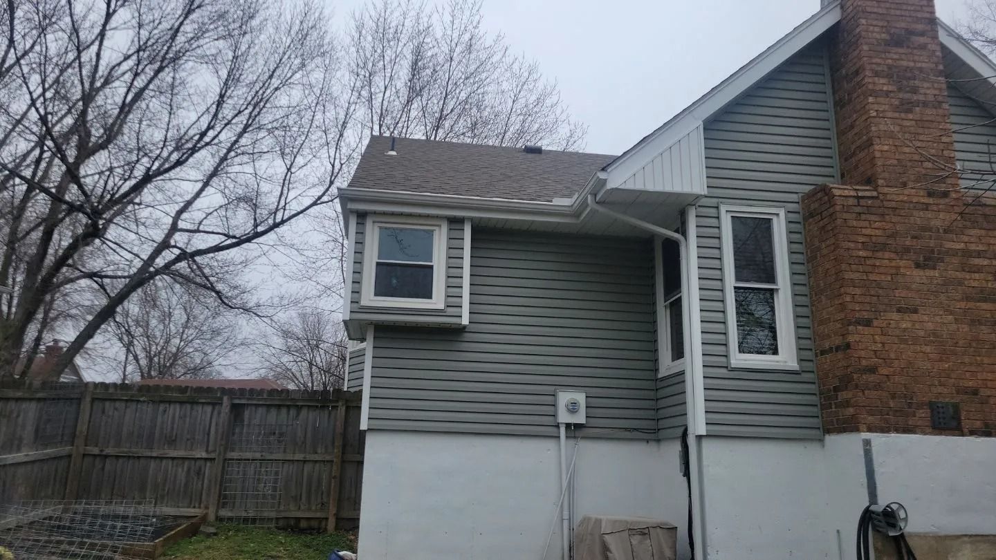 Side of a house with gray siding, two windows, and a brick chimney under a cloudy sky.