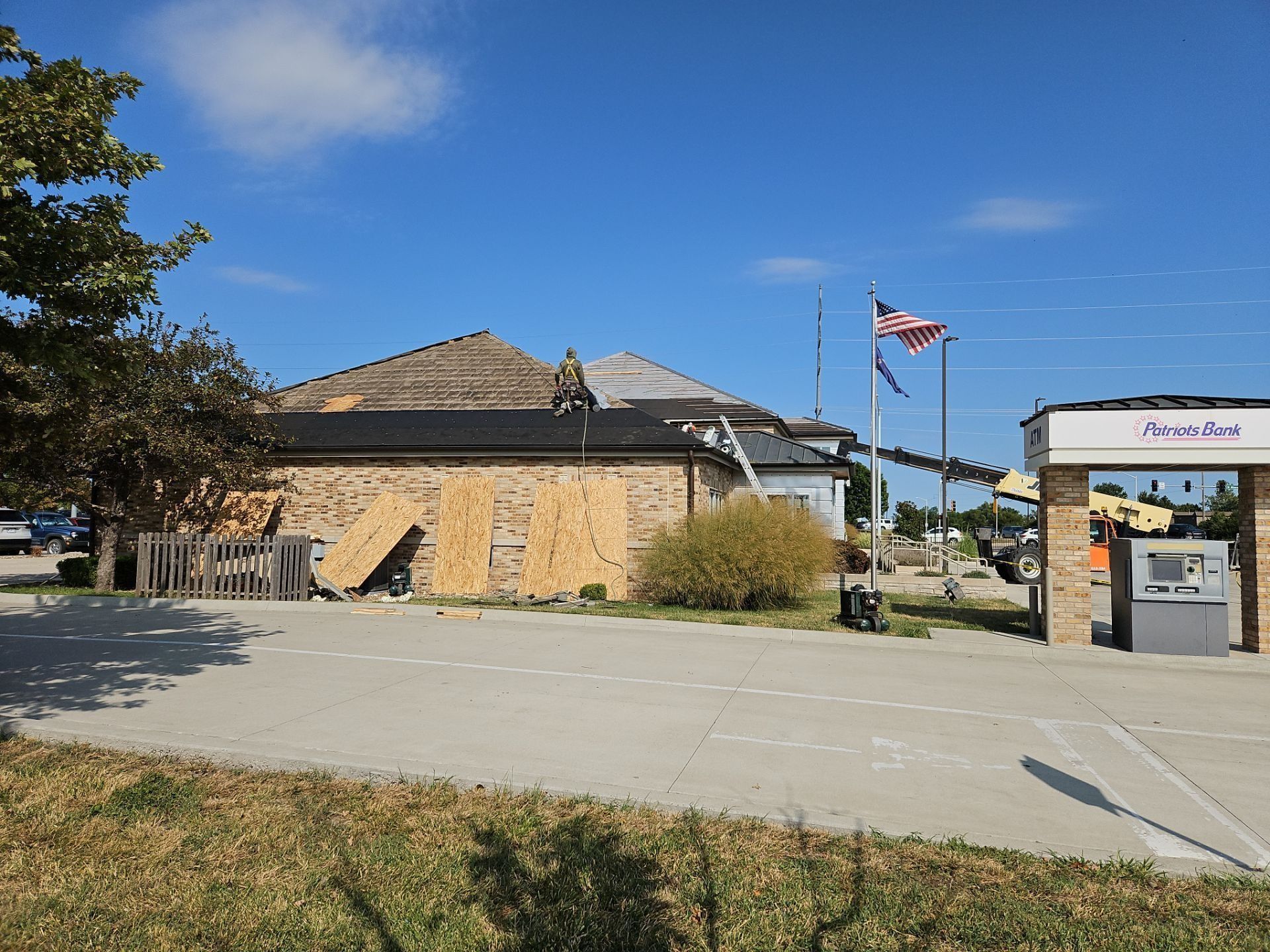Building with roof damage, boarded up siding, American flag, ATM, blue sky.