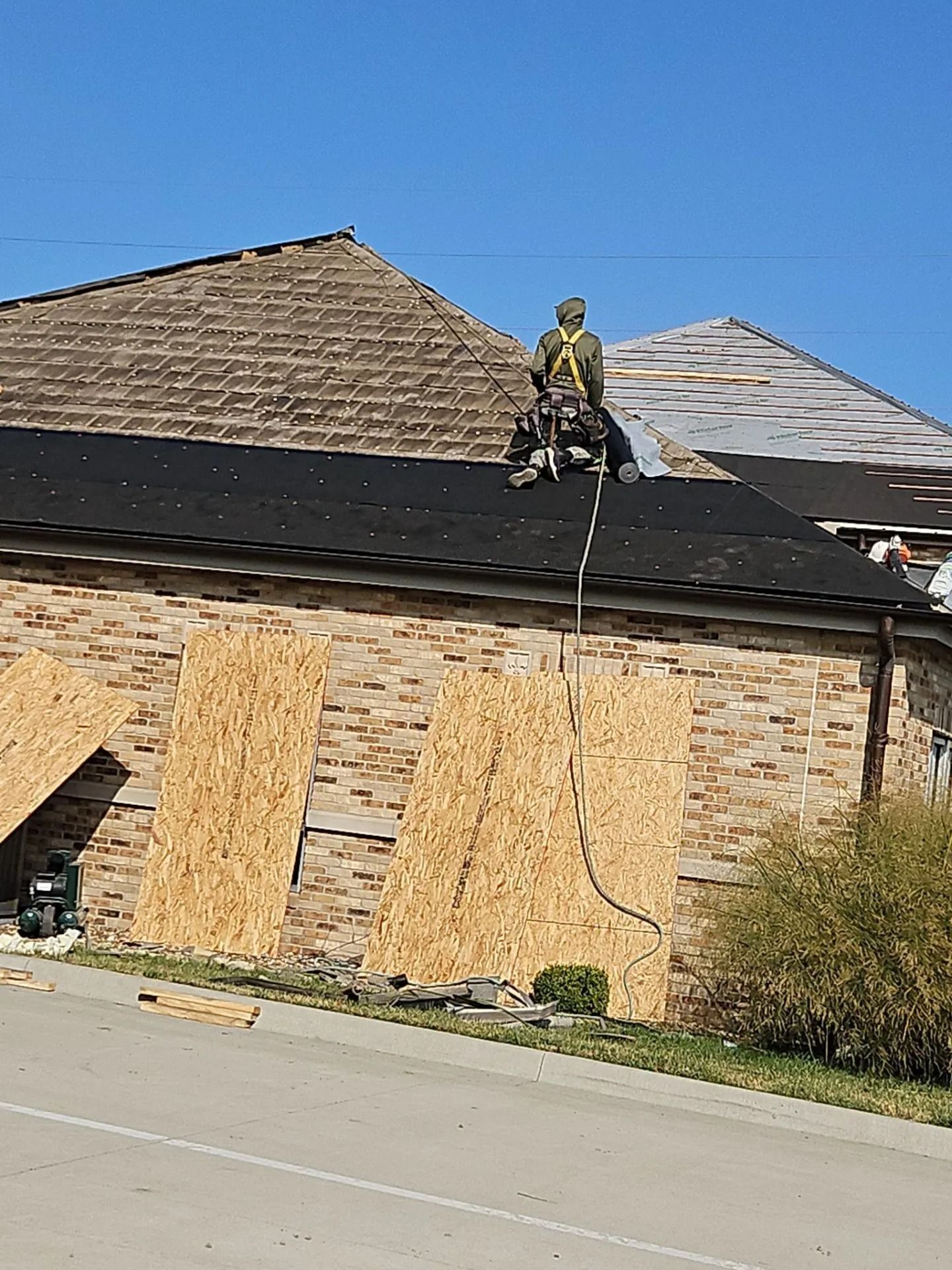 Roofer on a roof wearing safety harness, boarding up windows, and working near a street.