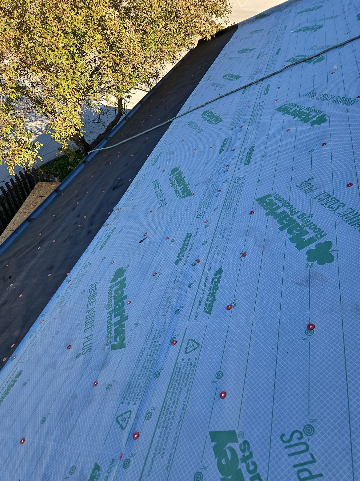 Rooftop with blue underlayment and green print, next to older roofing, with a tree in the background.