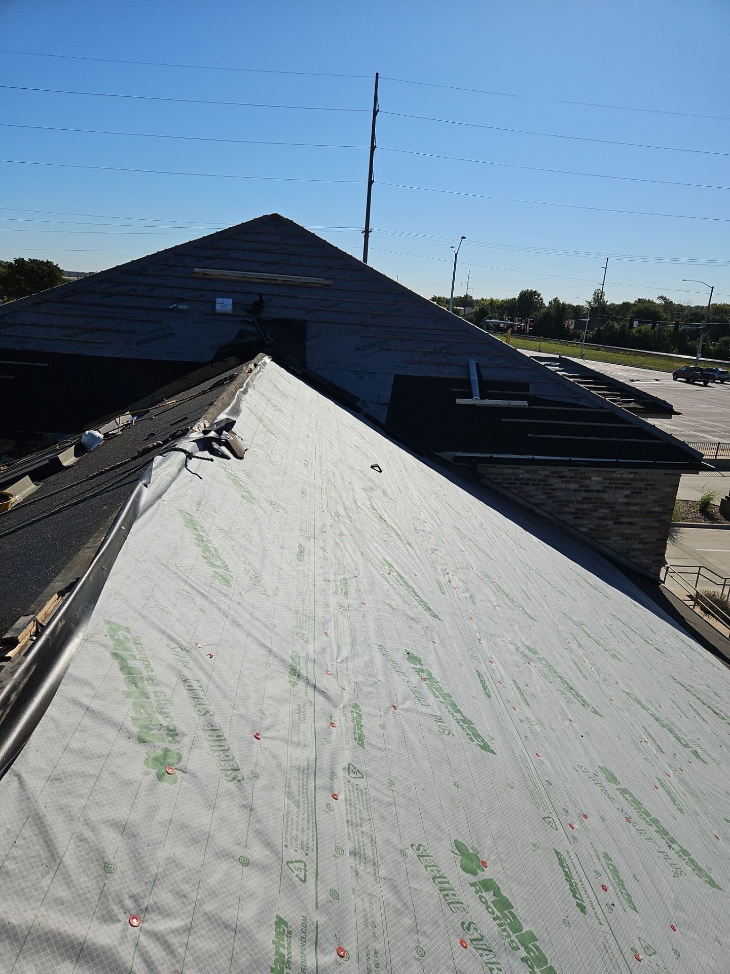 Roof with exposed underlayment and partially installed shingles. Blue sky and parking lot visible.