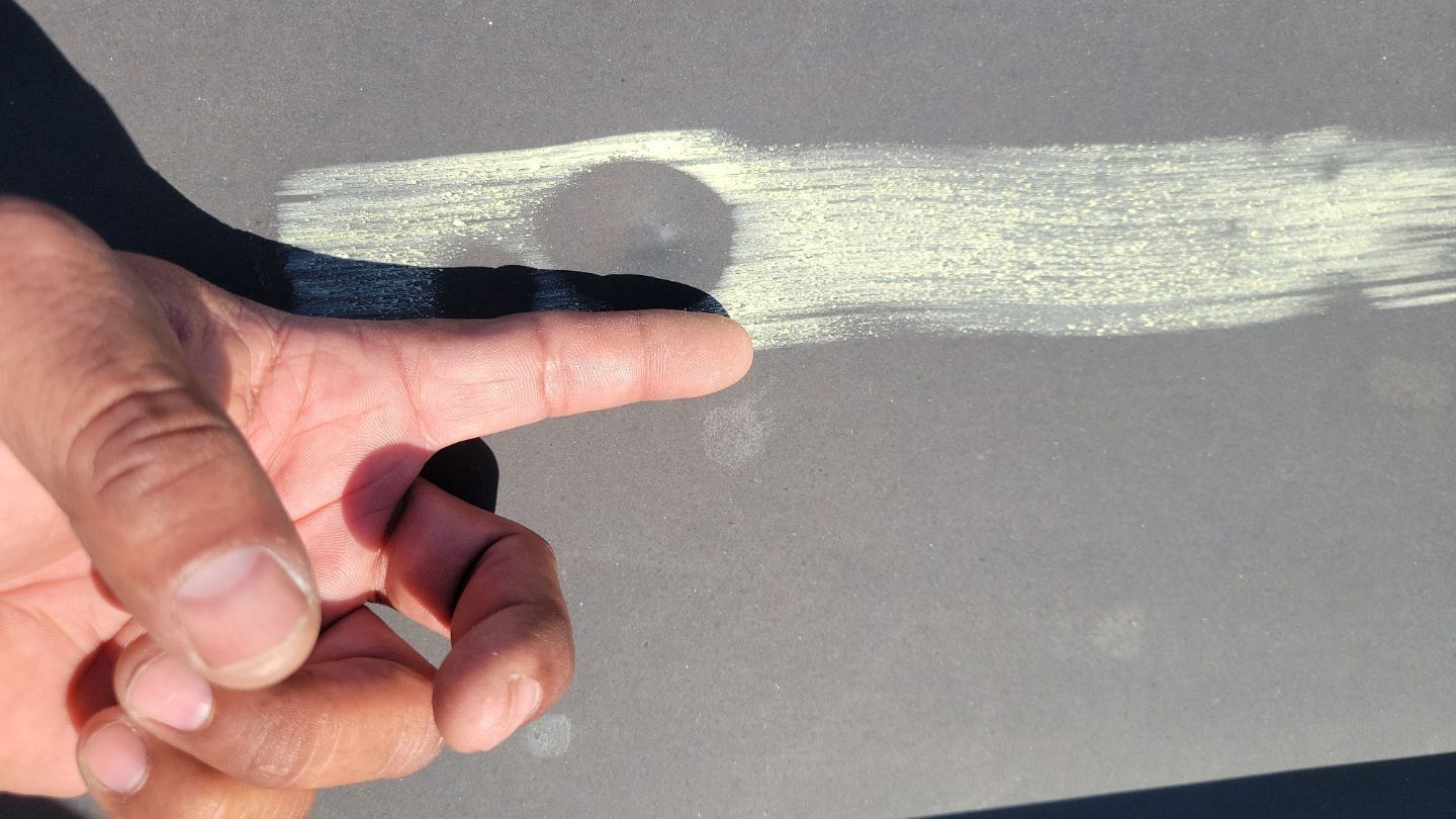Hand pointing to a scratch and circle on gray surface, possibly a car.