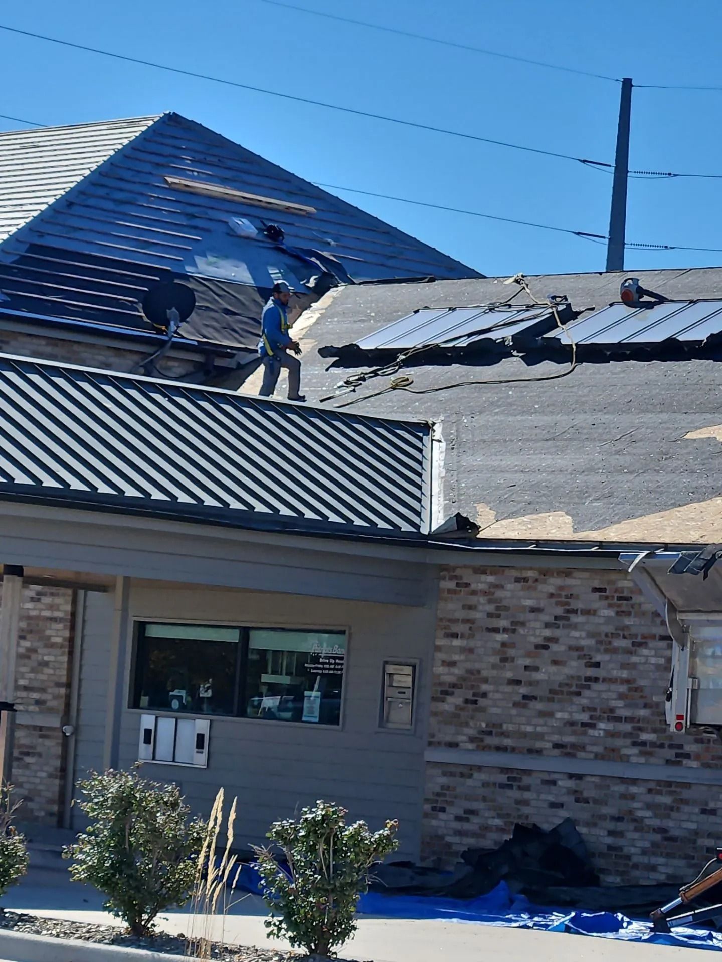 A person repairs a damaged roof, standing on a corrugated metal surface, blue sky overhead.