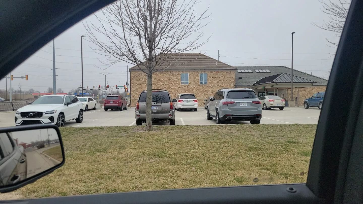 View from a car window of a parking lot with several vehicles parked in front of a brick building on a cloudy day.