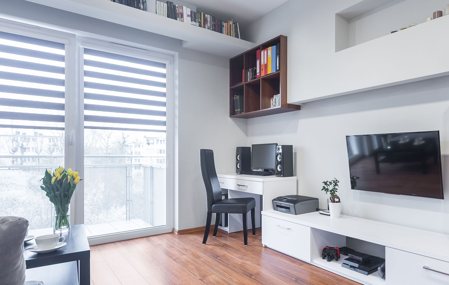A living room with hardwood floors , a desk , a television , and a balcony.