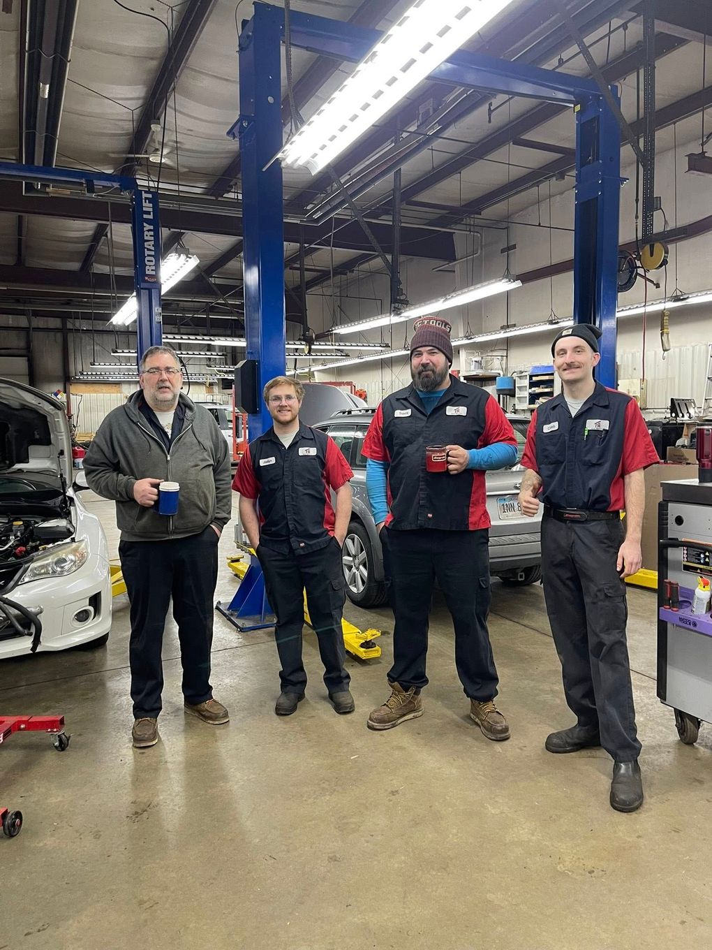 Four mechanics standing in an auto repair shop, holding mugs and smiling near vehicle lifts.