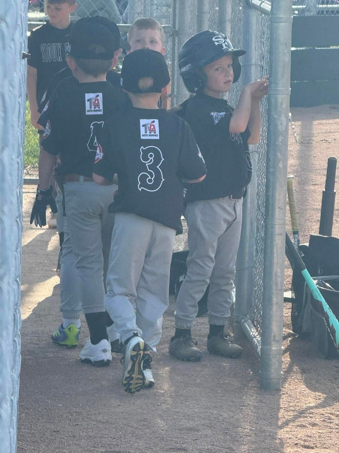 Kids in black jerseys with the T&A brand and gray pants walking along a chain-link fence at a sports field