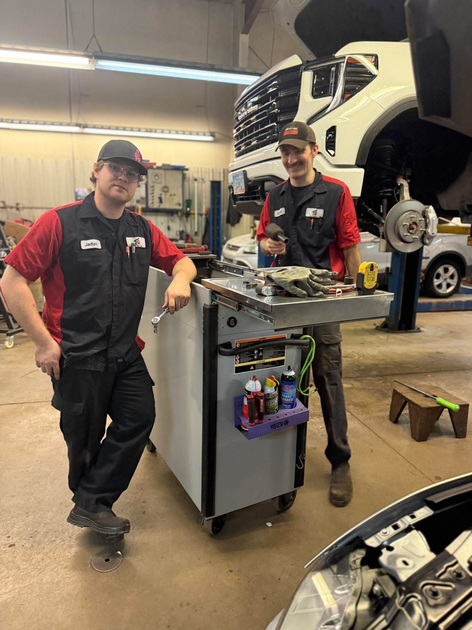 Two mechanics in red-and-black uniforms beside a tool cart in an auto repair shop, with a car on a lift behind them.