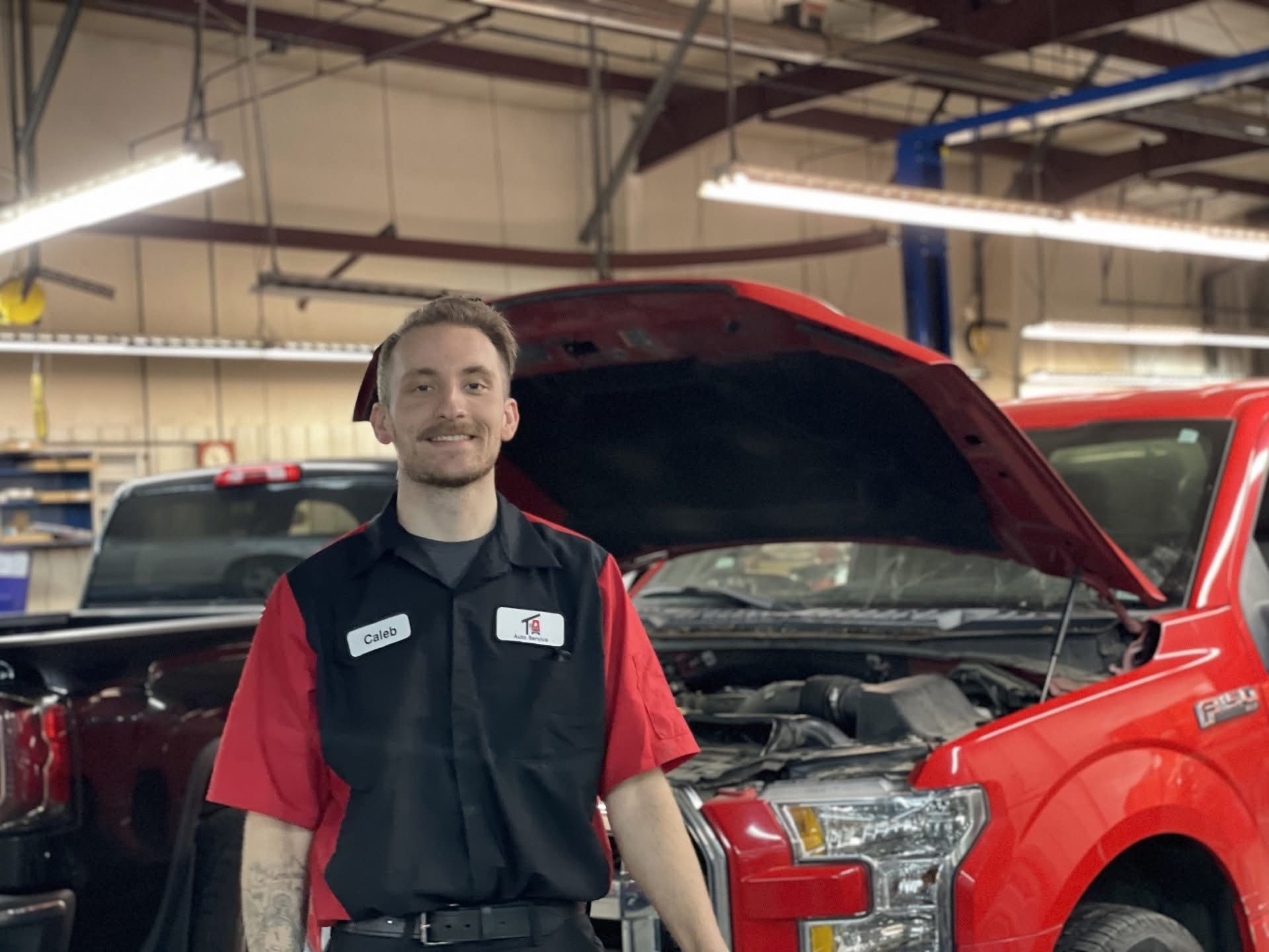 Mechanic in black and red uniform standing beside a red pickup truck with hood open in a garage