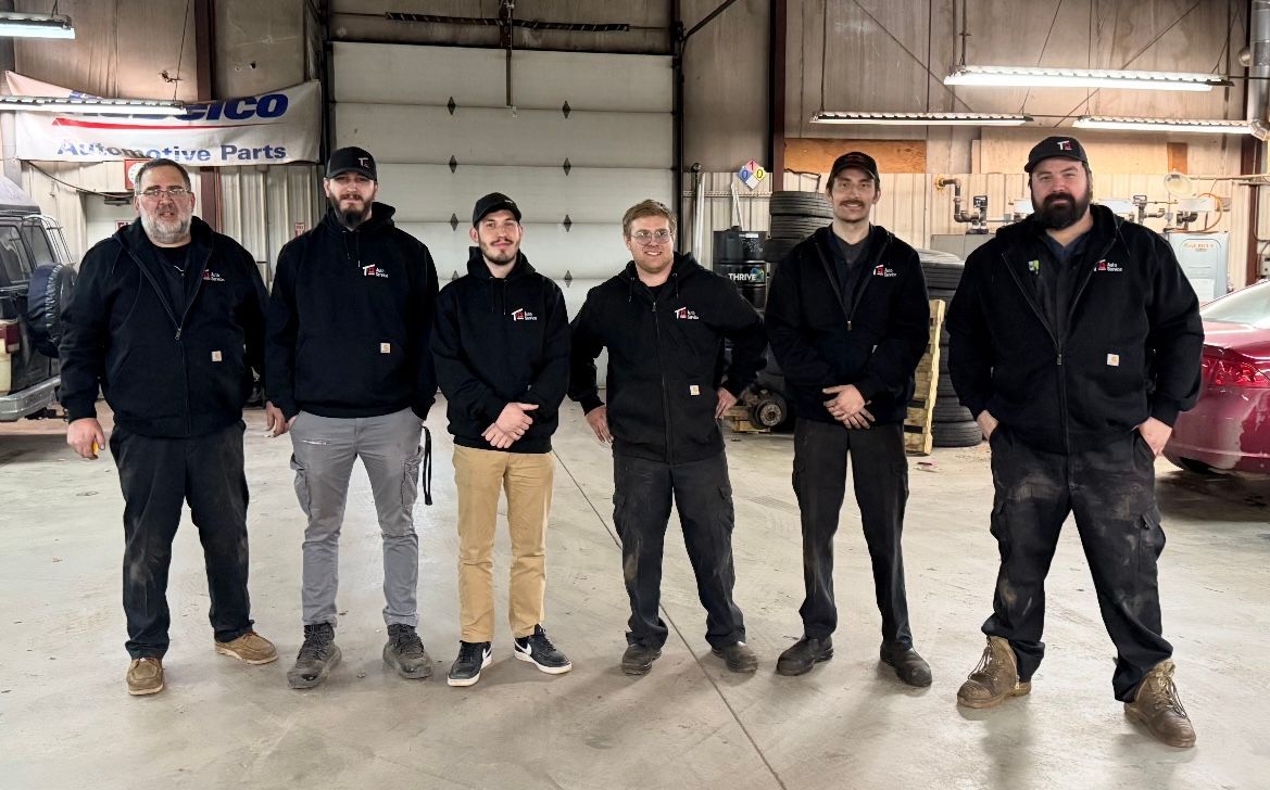 Six people in black work jackets stand in a warehouse with red machinery in the background.