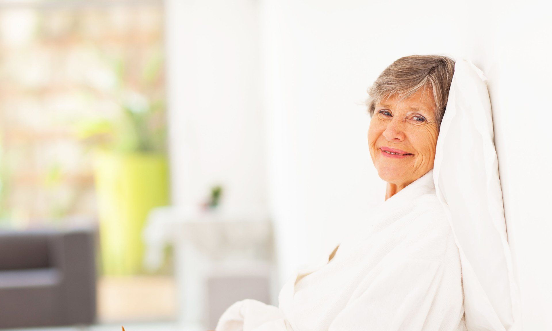 An elderly woman in a bathrobe is sitting on a couch.