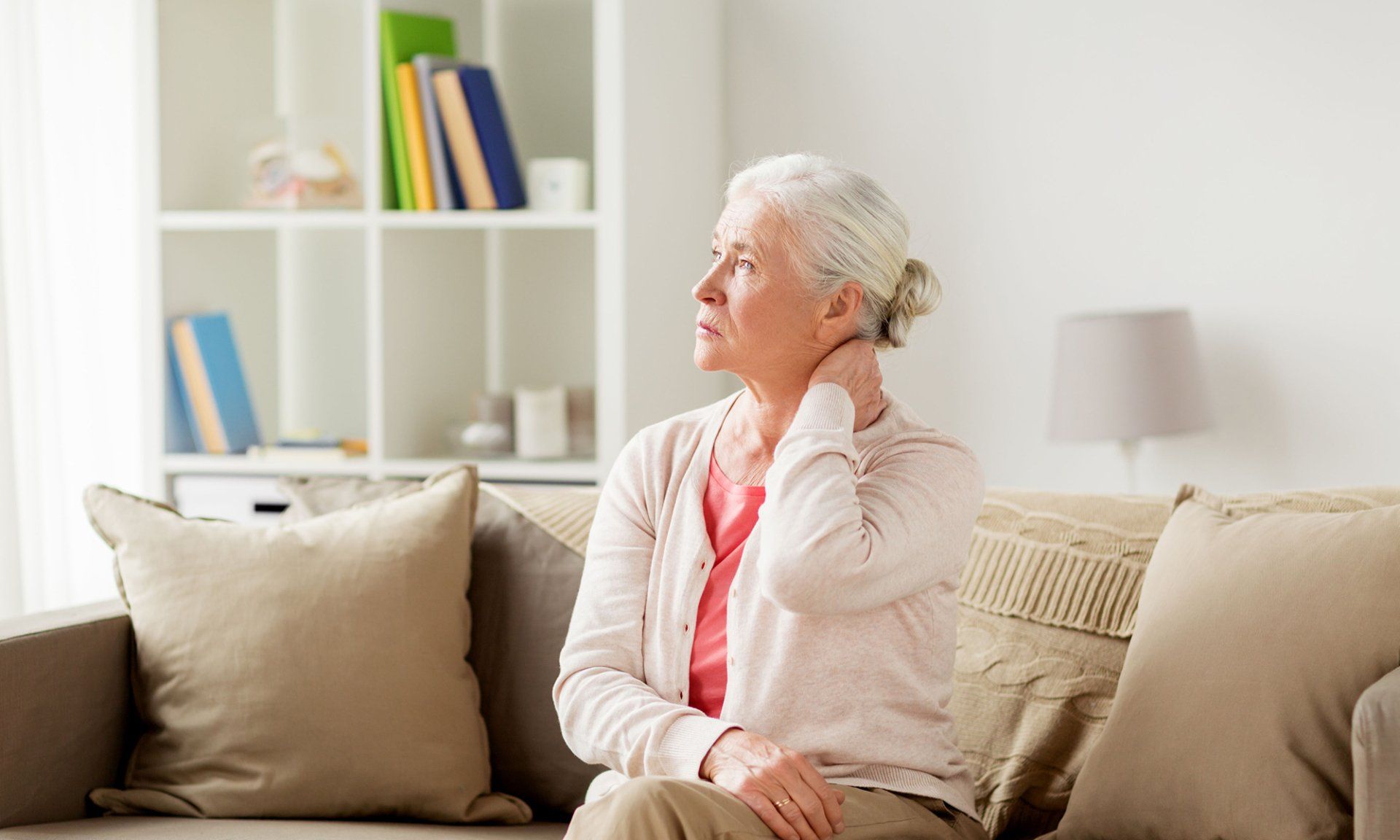 An elderly woman is sitting on a couch holding her neck in pain.