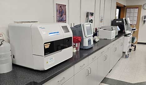 Laboratory countertop with a large white analyzer, smaller machines, and cabinets in a clinical workspace