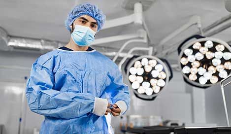 surgeon in blue scrubs, mask, and cap, in a bright operating room with surgical lights
