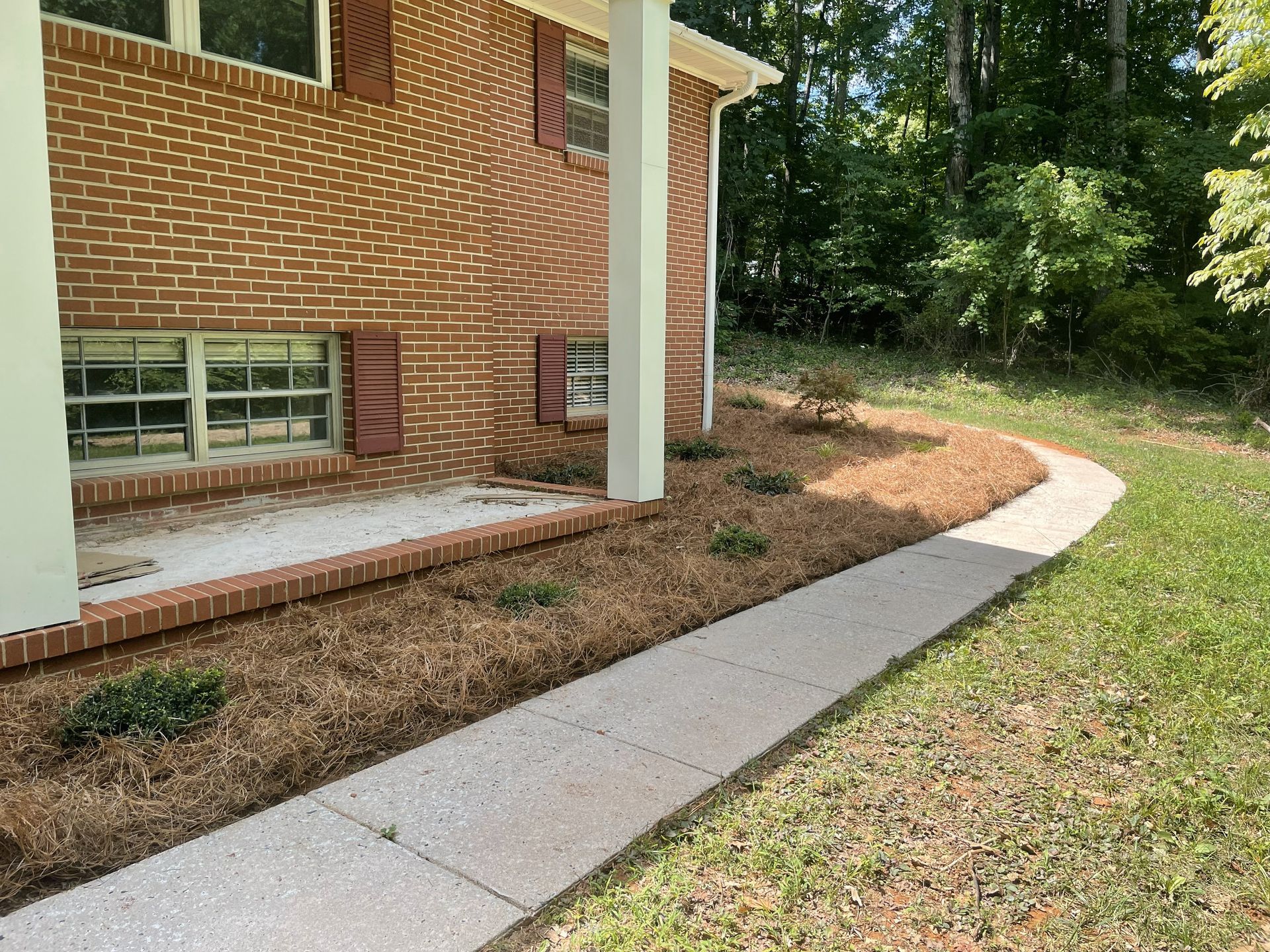 Brick house with a concrete walkway, mulch bed with greenery, and trees in the background.