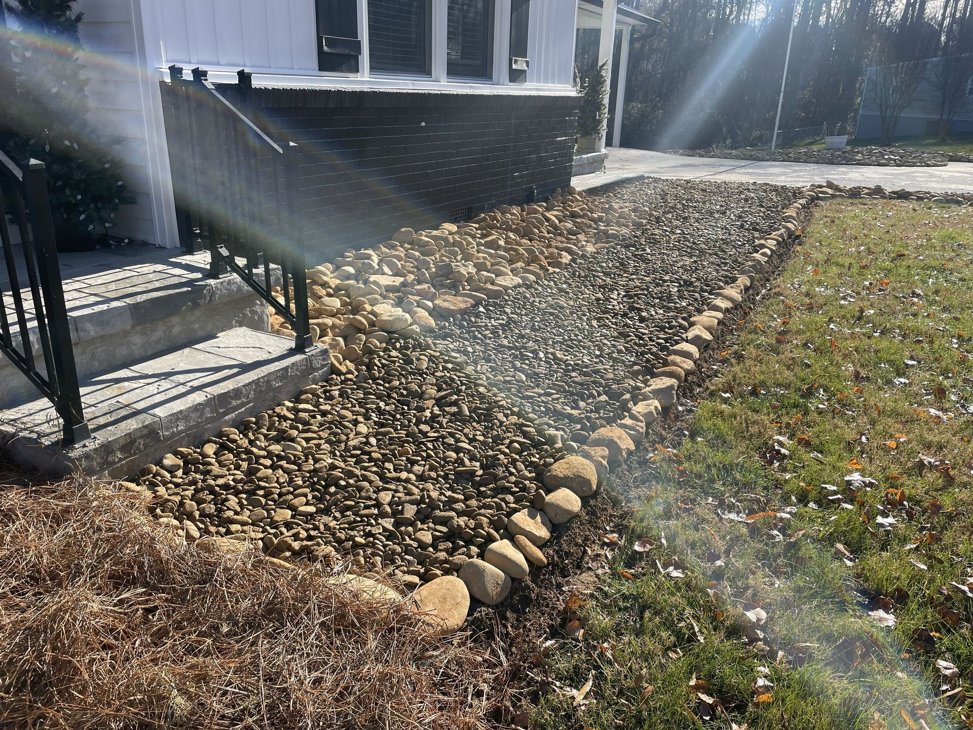 Rock-filled garden bed next to a house's steps and a grassy lawn, with sun shining.