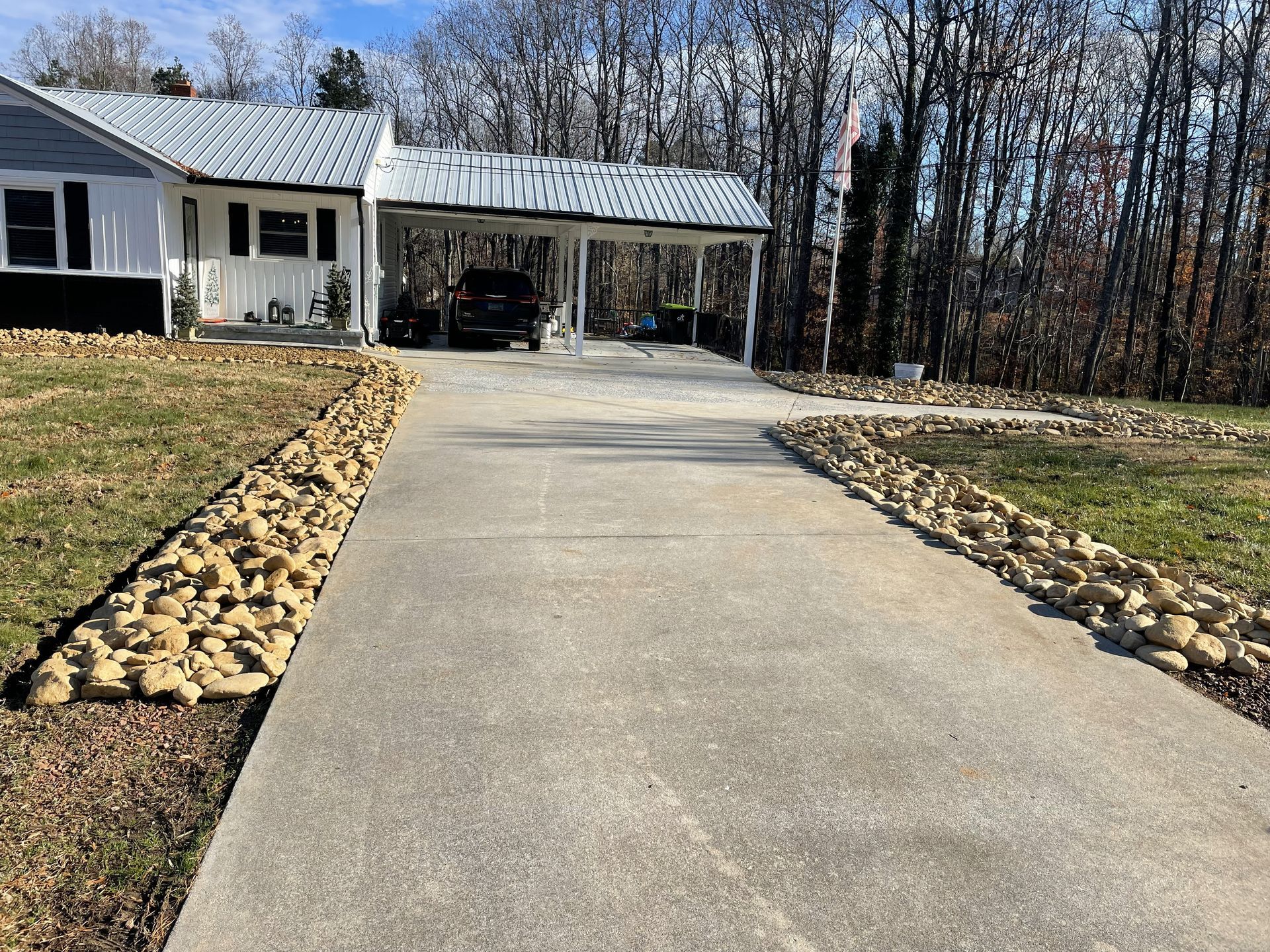 Concrete driveway lined with rocks leads to a carport and house. Bare trees are in the background.