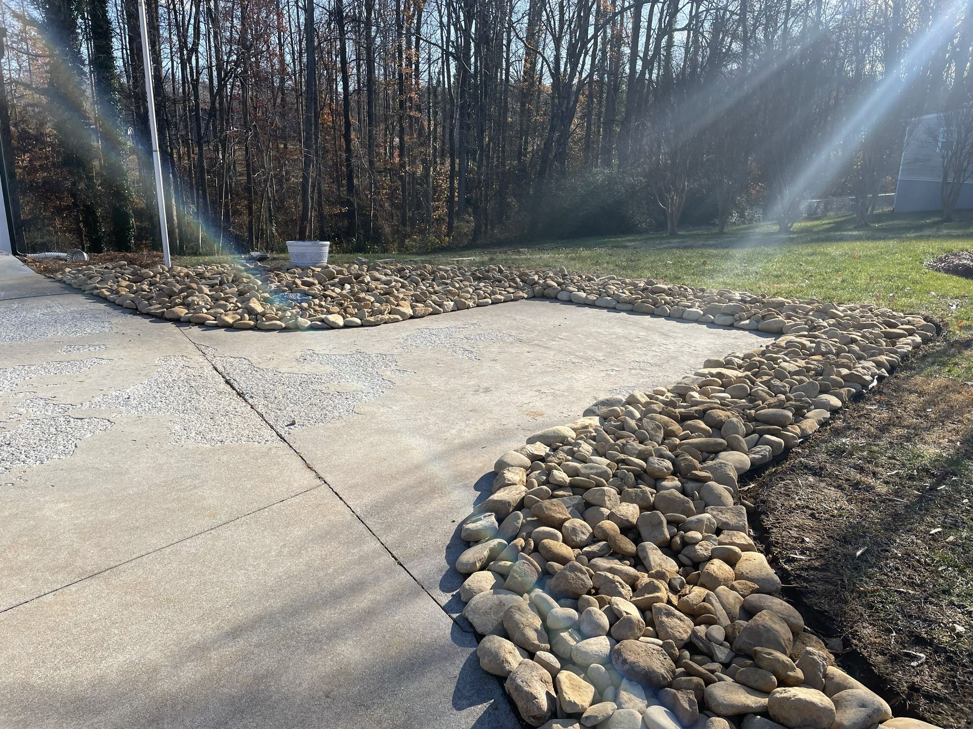 Concrete driveway bordered by rocks, leading to a grassy area with trees and sunlight.