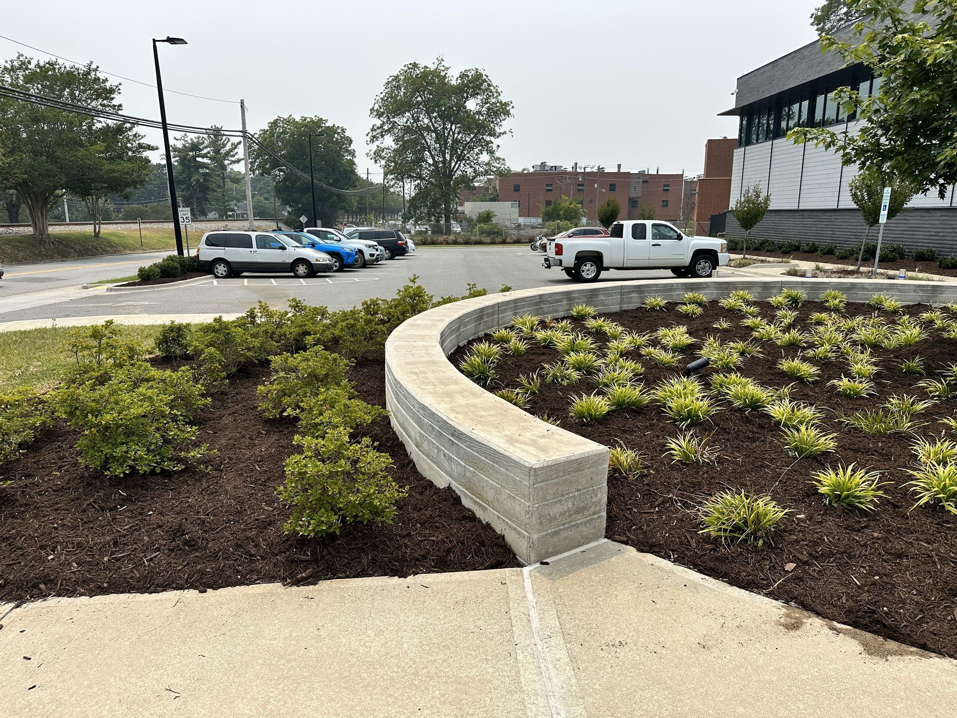 Curved concrete wall with landscaping. Cars parked in lot. White pickup truck drives by.