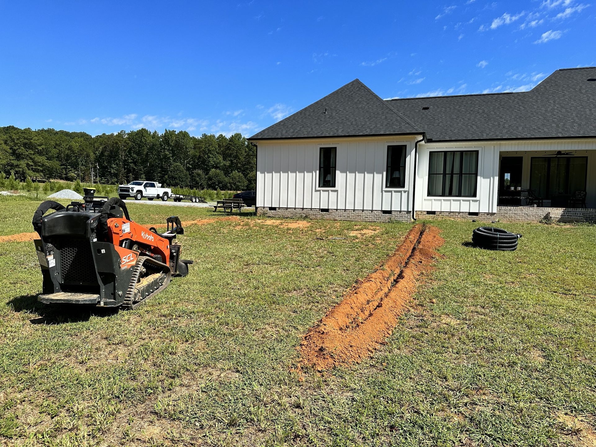 A small trenching machine digging a line in a grassy yard next to a white house under a blue sky.
