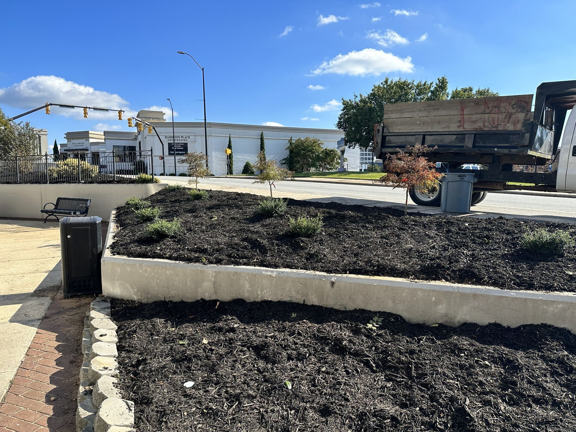 A flower bed with dark mulch, trash can, building and truck. Blue sky.