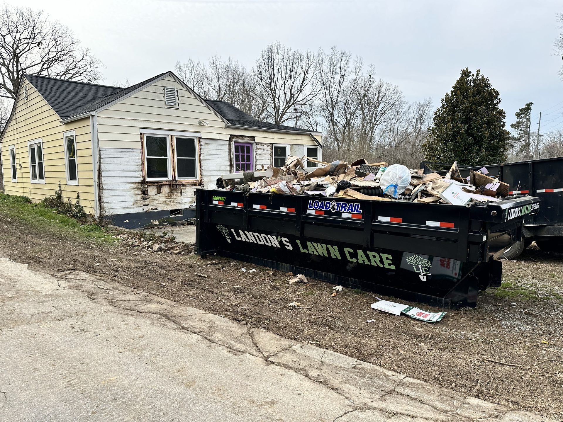 Dilapidated house with a dumpster overflowing with debris in front. Lawn care company logo is on the dumpster.