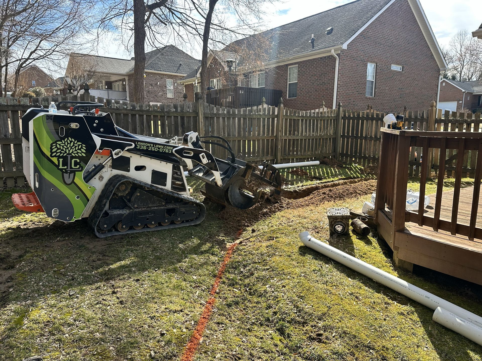 Mini excavator digging a trench in a backyard next to a wooden deck and a fence. White PVC pipes are nearby.