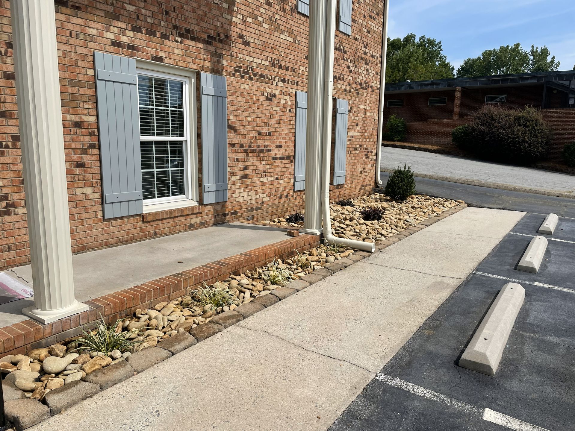 Brick building with window and grey shutters, next to a concrete walkway and parking area.