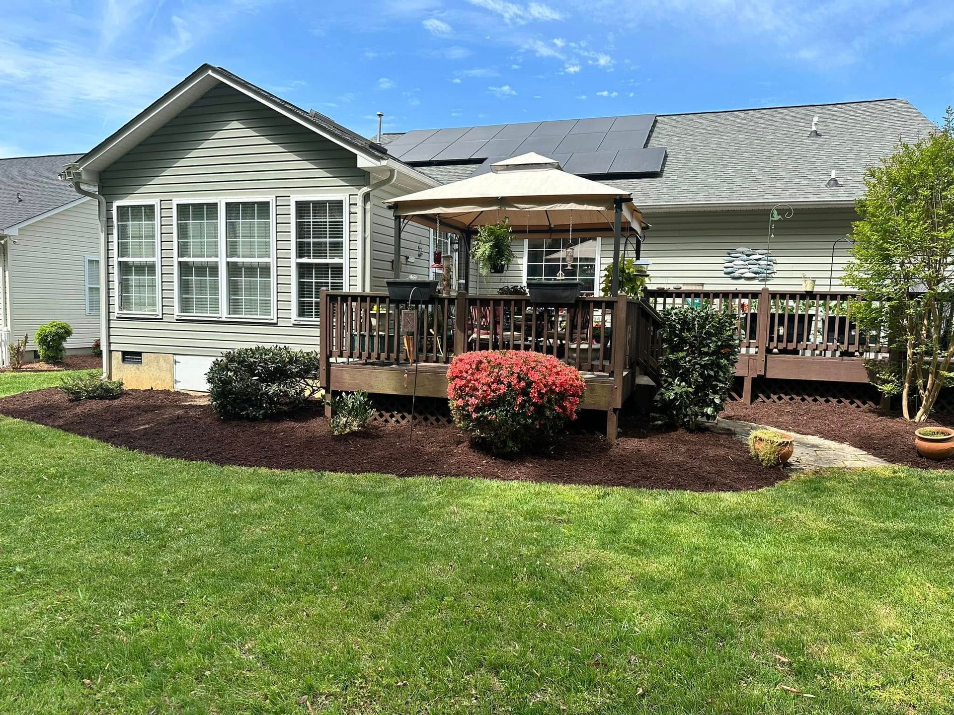 Backyard deck with gazebo, surrounded by landscaping and house with gray siding under a blue sky.