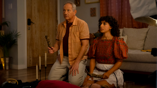 Two people sit on a living room floor watching something offscreen, one holding a remote.