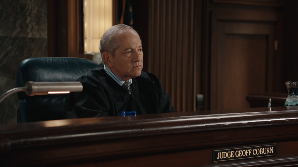 Judge sitting at a courtroom bench behind a nameplate, looking to the side, with a gavel nearby.