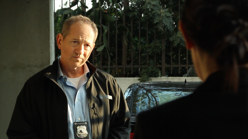 A man in a jacket and badge faces someone off-camera in a sunlit parking area.