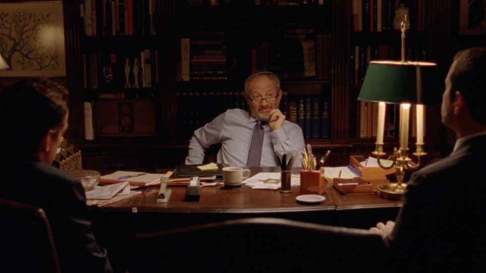 Dimly lit meeting room with three people seated around a desk, papers and lamps scattered across it.