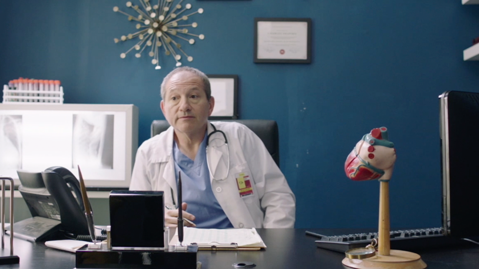 Doctor in white coat seated at a desk in a teal office, with a model heart on the right