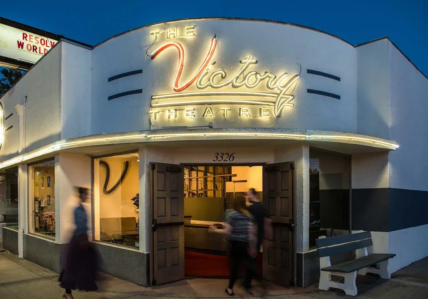 Neon-lit storefront with open doors and a person standing in the entrance at dusk.
