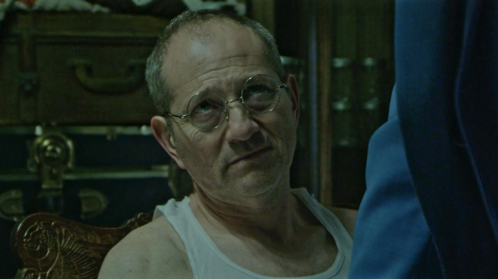 Man in glasses and white tank top looks up in a dim room, facing someone in blue.