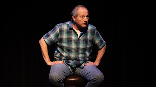 Man seated on a stool against a black background, hands on hips, wearing a plaid shirt and jeans.