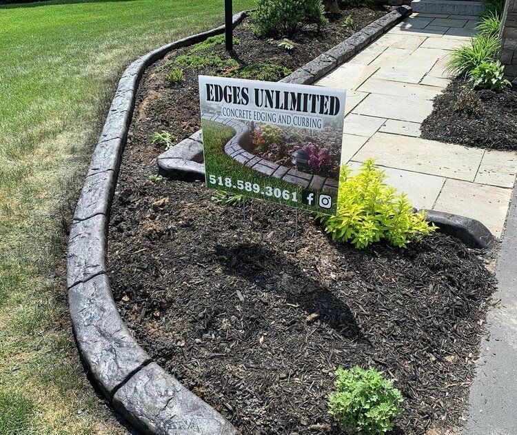 a sign for edges unlimited is sitting in the middle of a garden next to a sidewalk