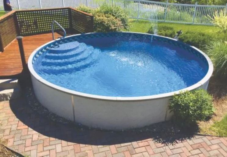 A circular above-ground swimming pool with built-in stairs, next to a wooden deck and a brick patio in a backyard.