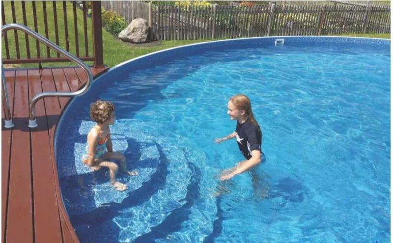 Two people play in a blue, round above-ground pool with a wooden deck and railing.