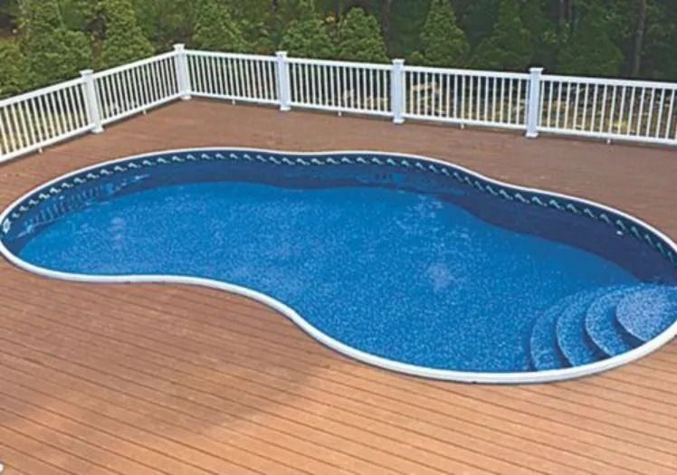 A kidney-shaped swimming pool with blue tiled steps, surrounded by a wooden deck and a white fence in a backyard.