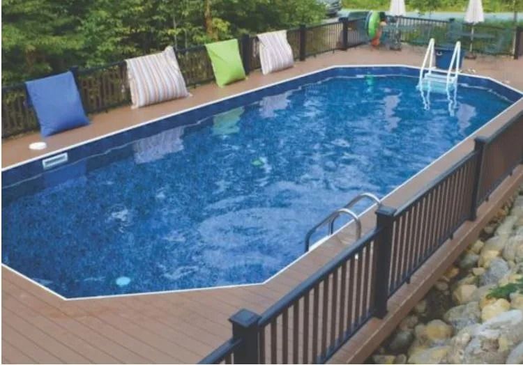 An oval above-ground swimming pool with blue water and surrounding wooden decking, featuring railings and colorful pillows.