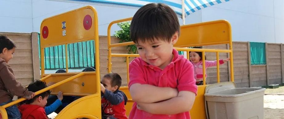 Child with arms crossed, appears upset, near yellow playground structure. Other children play nearby.