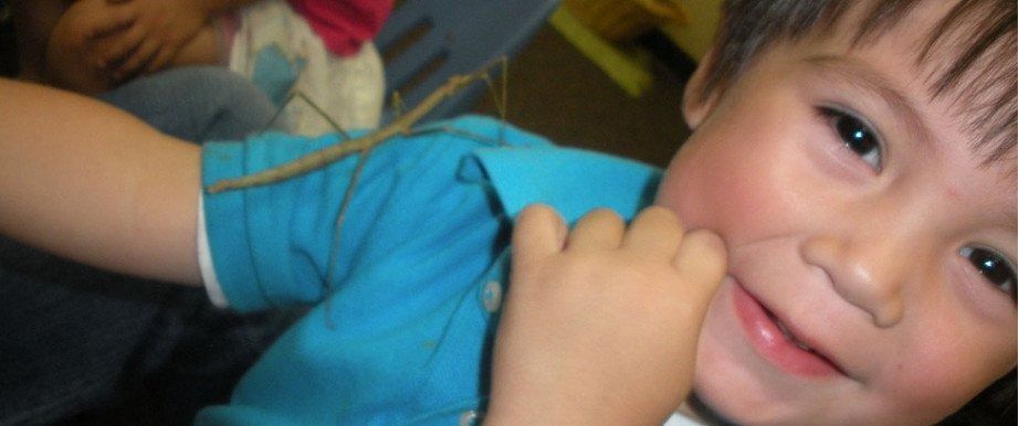 A young child in a blue shirt smiles, holding their hand up to their cheek.