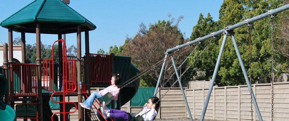 Two people on swings at a playground; one swings high, with a climbing structure and trees in background.