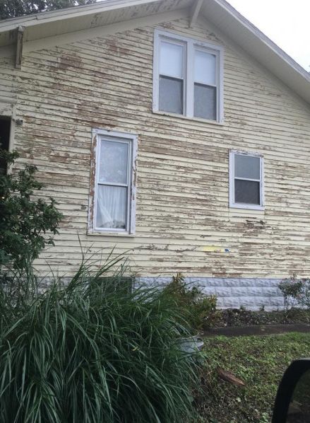 A weathered, two-story house with peeling yellow paint and three windows. Green foliage in the foreground.