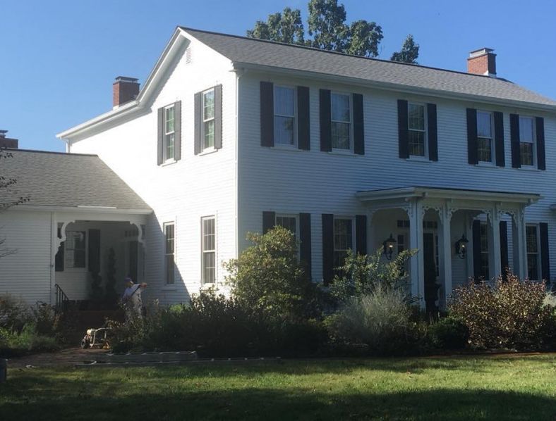 White two-story house with black shutters, brick chimneys, and a porch, set on a green lawn with shrubs.